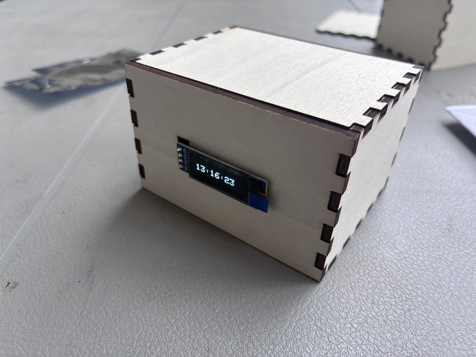 A laser-cut plywood box with finger joints, an OLED display in the front panel showing the time 13:16:23