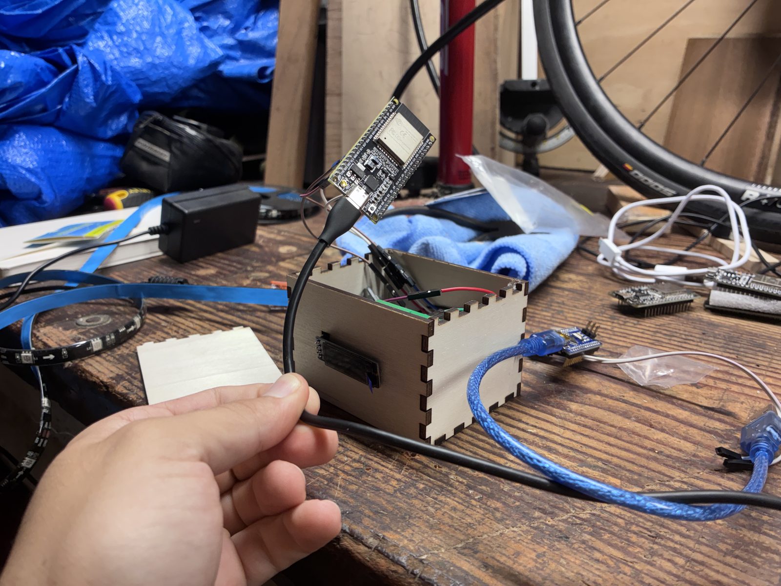An ESP32 development board wired by jumper leads into the opened plywood box, on a cluttered workbench